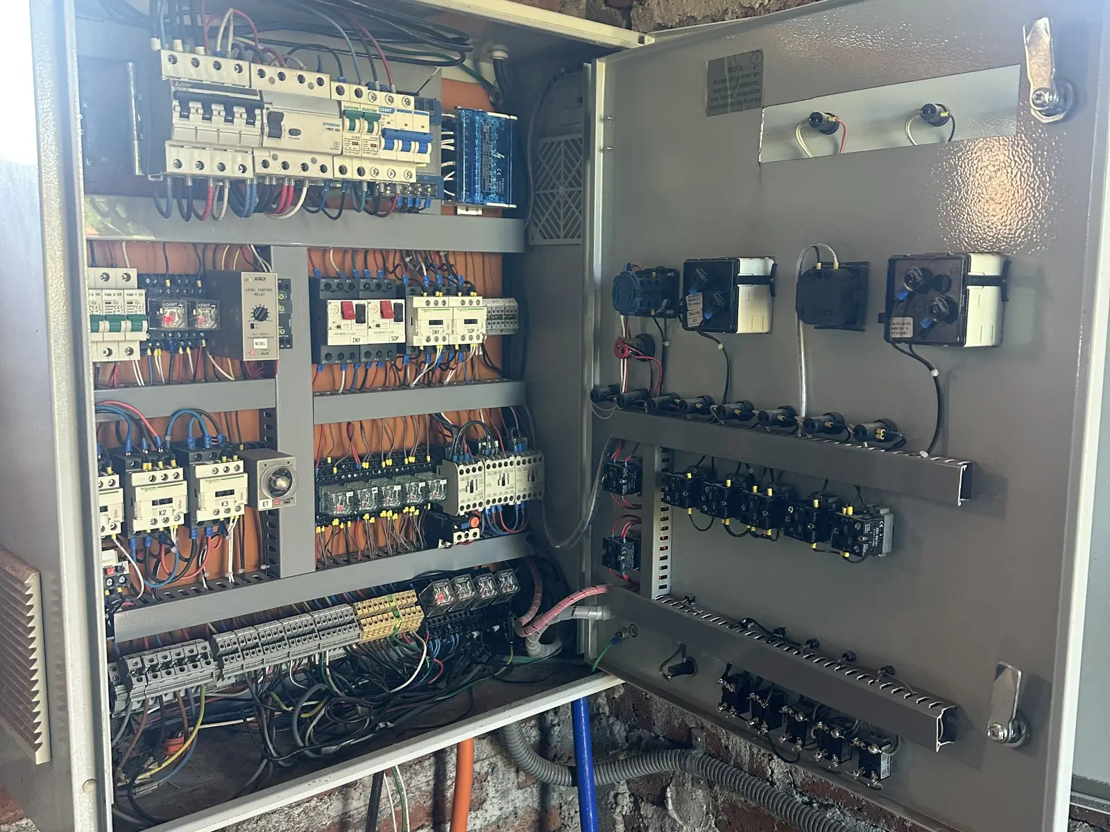 Interior de tablero eléctrico abierto con cableado y breakers sobre pared de ladrillo.